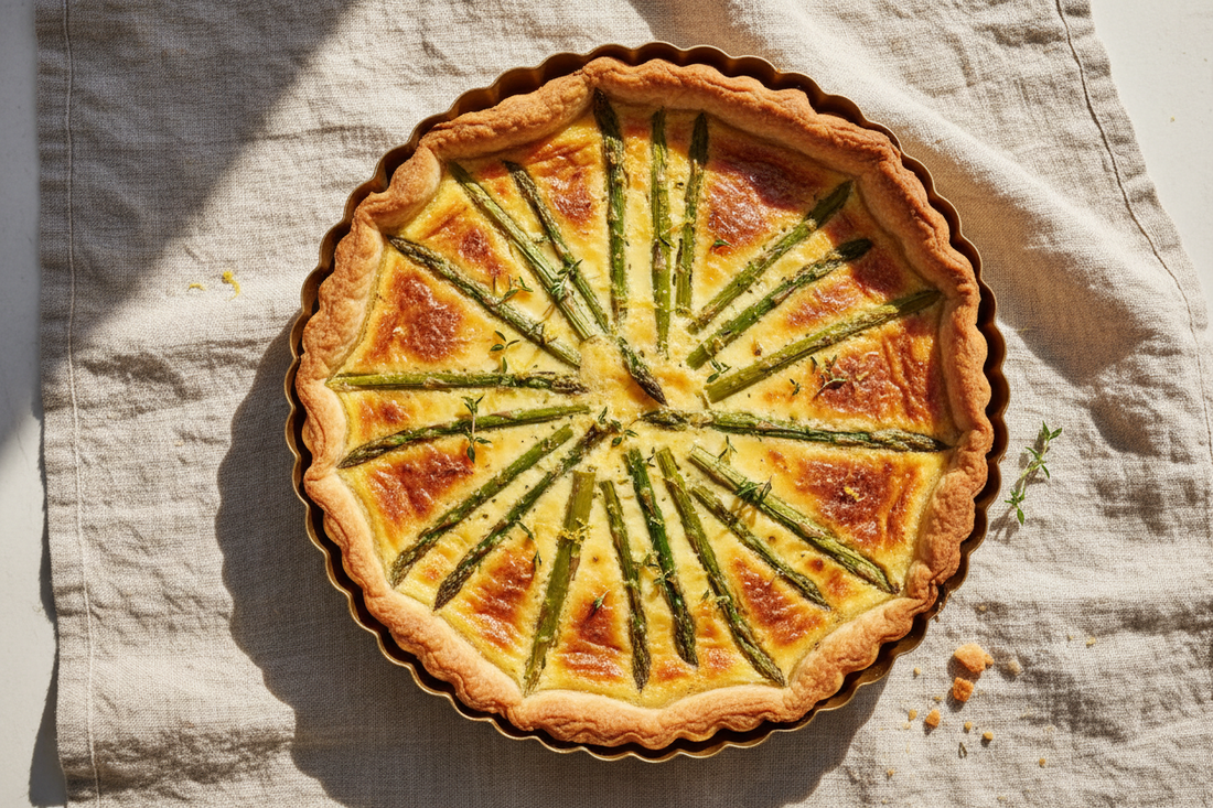 overhead shot of the tart in a fluted tin with asparagus spears arranged across the golden Gruyère custard, lemon zest and thyme scattered alongside.