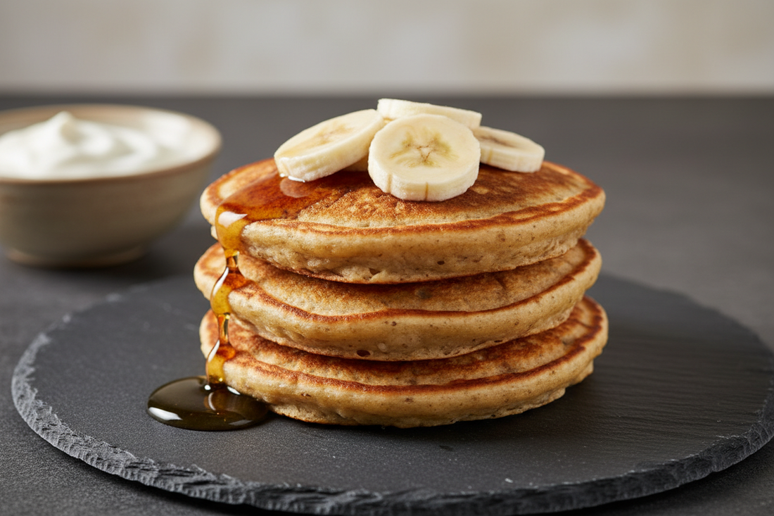  small stack of golden banana-oat pancakes on a dark slate plate, topped with fresh banana slices and a drizzle of maple syrup, shot at a 45-degree angle with soft natural side light