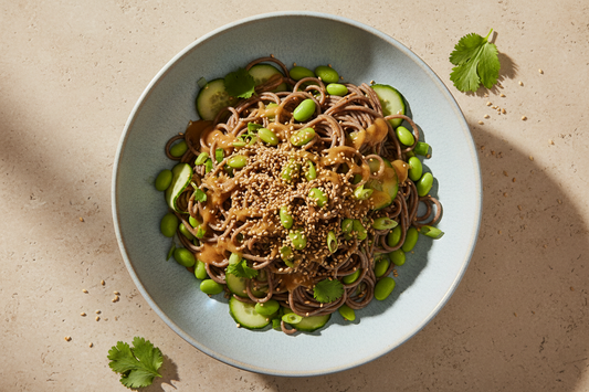 Overhead shot of a chilled sesame soba noodle salad in a ceramic bowl with edamame, cucumber, green onions, fresh cilantro, and toasted sesame seeds on a linen surface.
