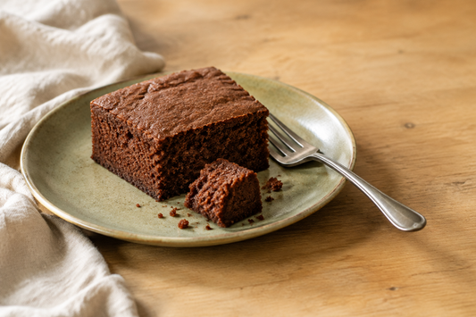 A square of unfrosted chocolate cake on a sage-green plate, with a small bite separated to show the chocolate crumb