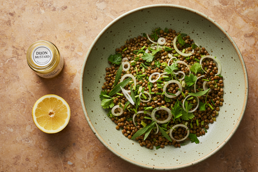 Overhead shot of the dressed Puy lentil salad with tarragon, parsley, chives, and shallot rings, Dijon jar and lemon at the edge of frame on a warm stone surface