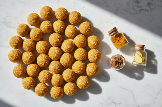 Overhead view of golden spice bliss balls coated in turmeric coconut sugar arranged on white marble, with small jars of turmeric, cinnamon, and ginger alongside.