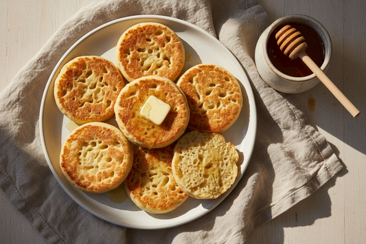 Overhead flat lay with golden crumpets, melting butter pooling into the holes, a honey pot with wooden dipper, linen napkin, and warm editorial lighting in your earthy neutral palette.