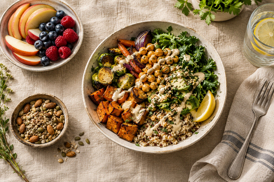 Overhead view of a bowl with roasted vegetables, chickpeas, greens, grains and creamy dressing, alongside sliced apples, berries, nuts and seeds.