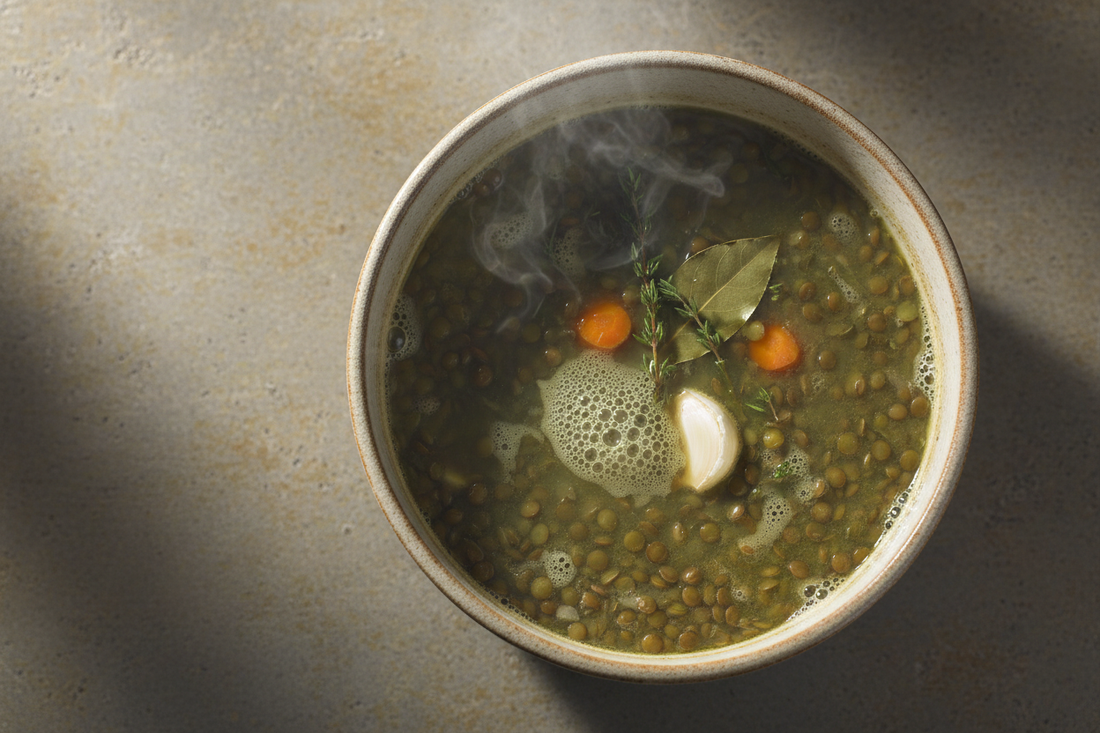 Overhead shot of a wide ceramic pot of gently simmering green lentils with bay leaf, garlic, thyme, and carrot visible — steam rising, warm natural light.