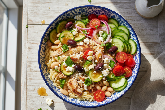 Overhead view of Mediterranean grain bowl with orzo, shredded chicken, white beans, sautéed zucchini, fresh cucumber, cherry tomatoes, kalamata olives, and crumbled feta cheese drizzled with honey-lemon dressing and garnished with fresh parsley