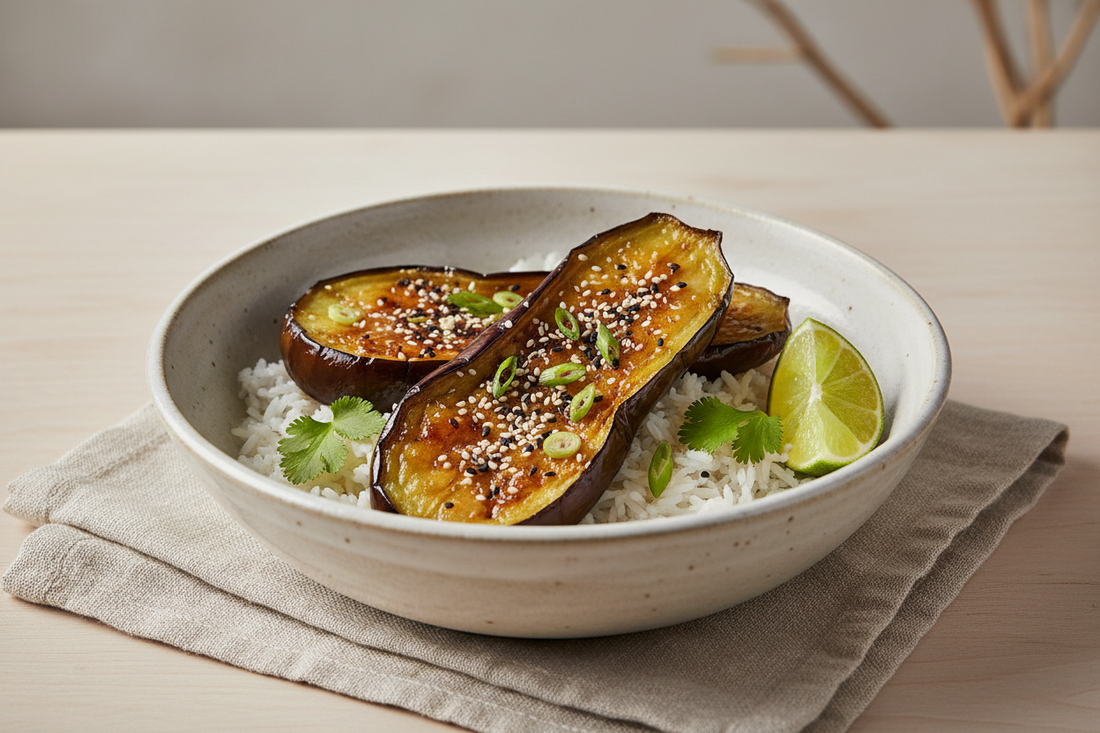 Miso-glazed eggplant halves served over sesame rice in a ceramic bowl, garnished with sesame seeds, green onions, cilantro, and a lime wedge.