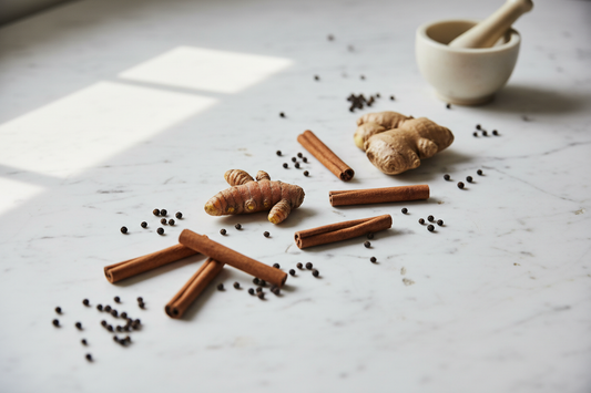 Whole turmeric root, fresh ginger, cinnamon sticks, and black peppercorns arranged on a marble surface with a ceramic mortar and pestle — the four foundational spices of The Grounded Kitchen's Spice Apothecary module.