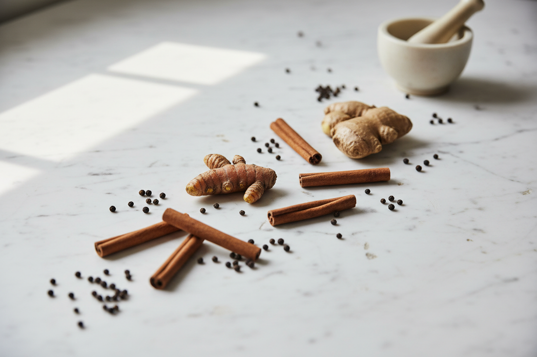 Whole turmeric root, fresh ginger, cinnamon sticks, and black peppercorns arranged on a marble surface with a ceramic mortar and pestle — the four foundational spices of The Grounded Kitchen's Spice Apothecary