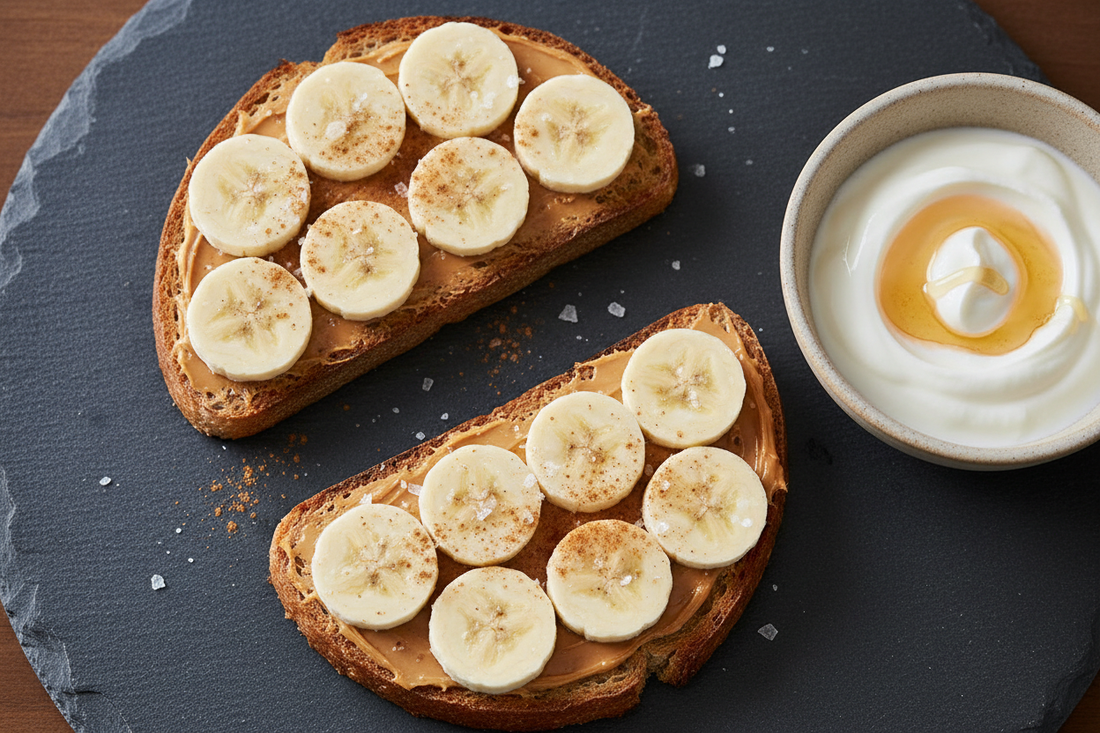 Two slices of golden sourdough toast spread with peanut butter and topped with banana slices, flaky salt, and cinnamon, served alongside a small bowl of yogurt with honey, on a dark slate surface shot at a 45-degree angle with soft natural side light