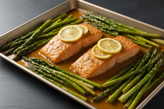 Two maple-lemon glazed salmon fillets with bright green asparagus and lemon rounds on a sheet pan, with caramelised pan juices, on a dark slate surface