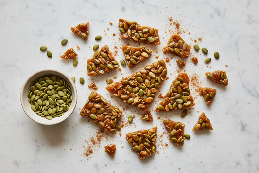 verhead view of spiced pumpkin seed brittle broken into irregular amber shards on white marble, with whole pepitas visible throughout and small ceramic bowl of pumpkin seeds alongside.